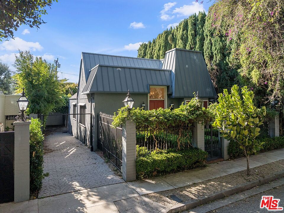 A 1925 cottage with steep gables, a standing-seam metal roof, and a black iron fence draped in greenery, captured in golden afternoon light.